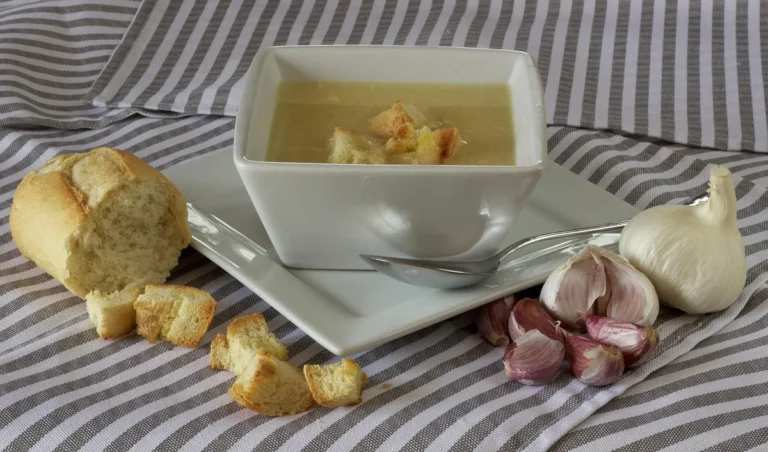 Miska česnekové polévky s krutony, vedle česnek a pečivo.