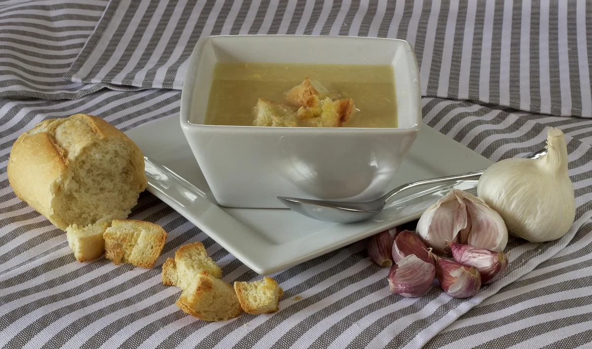 Miska česnekové polévky s krutony, vedle česnek a pečivo.